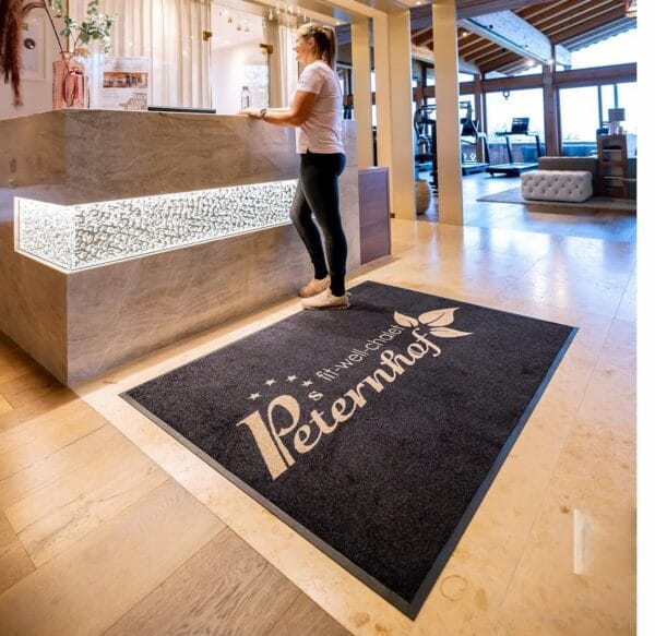Tapete de entrada personalizado com logótipo dourado, aplicado numa receção de hotel com zona de fitness ao fundo
